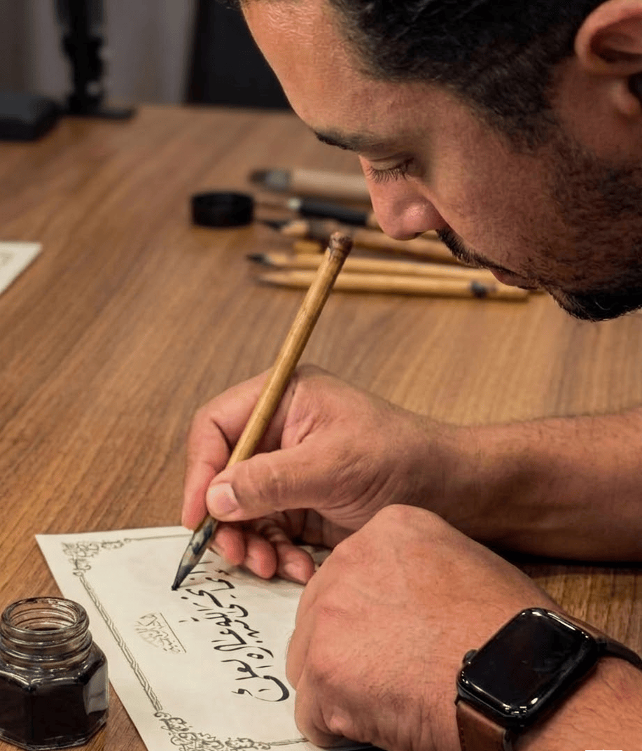 A professional close-up photograph of Mohamed Bakr in profile, deeply focused on performing traditional Arabic calligraphy with a reed pen (Qalam) and black ink. He is writing the Quranic verse: 'إنما يخشى الله من عباده العلماء' ('Those truly fear Allah, among His Servants, who have knowledge' — Fatir:28) in Ruq'ah script. The photo also features a traditional ink well positioned on the parchment.
